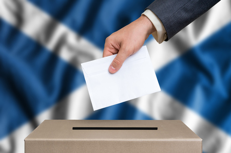 Election in Scotland. The hand of man putting his vote in the ballot box. Scottish flag on background.の写真素材