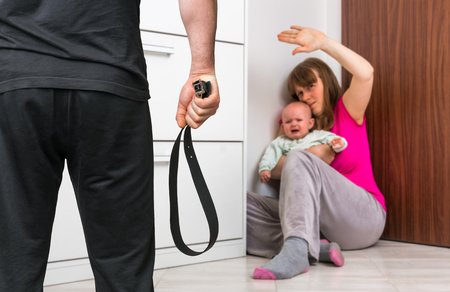 Aggressive man with a leather belt is abusing his wife and crying baby, woman covering in the corner at home - domestic violence conceptの写真素材