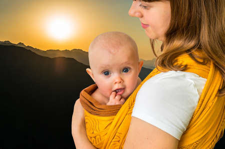 Happy mother with her baby in a sling carrying him in mountains during sunsetの写真素材