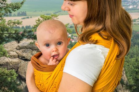 Happy mother with her baby in a sling carrying him in parkの写真素材