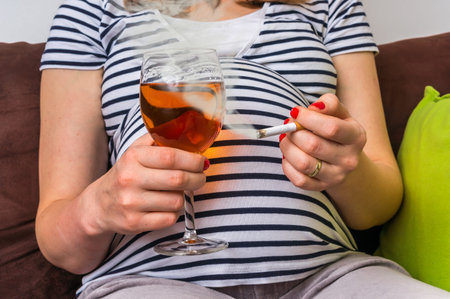 Young pregnant woman holding wine and cigarette at homeの写真素材