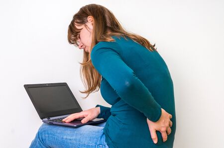 Woman is suffering from back pain sitting with laptop on white background - concept of bad posture and bad sitting positionの写真素材