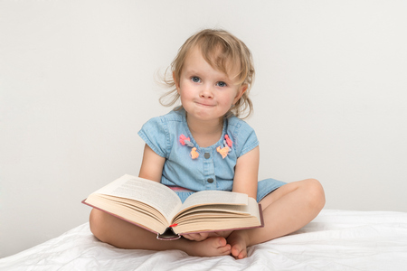 Cute little child in blue dress reading a book and sitting on bedの写真素材