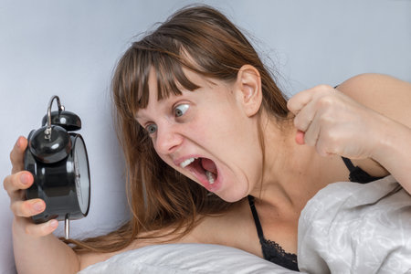 Shocked woman with alarm clock comes late to work because she oversleep - oversleeping conceptの写真素材
