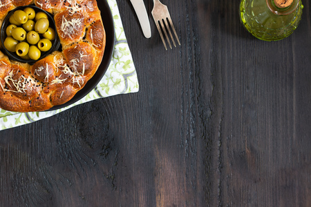 Savory pie, round monkey bread with stuffed buns oives and cheese, topped with grated cheese and black sesame bowl pitted green olives in center on black table with fork and knife olive oil copy spaceの写真素材