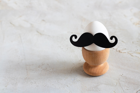 white egg with black moustache in wooden egg cup on grey cement textured background with copy space, concept for breakfast and easterの写真素材