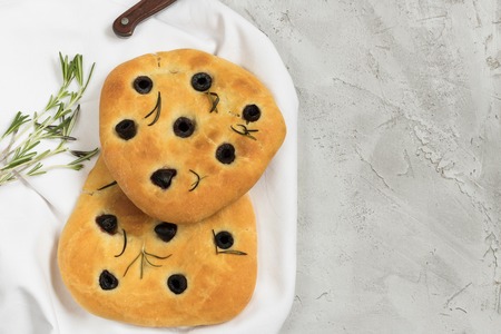 Traditional Italian Focaccia with black olives and rosemary - homemade flat bread focaccia, two baked pices on white table cloth and grey concrete table withknife handle and fresh rosemaryの写真素材