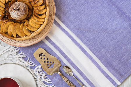 Home made apple pie topped with apple in pastry cover in left upper corner of horizontal shot. cup of red berry tea, cake server, spoon on the violet table cloth on light grey wooden textured table. Copy spaceの写真素材