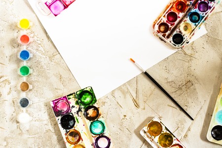 Flatlay with bright water colour paints in small round containers and palettes as frame, small brush and white empty sheet of paper in center on grey cement backcground, mess on artist desk, with copy spaceの写真素材
