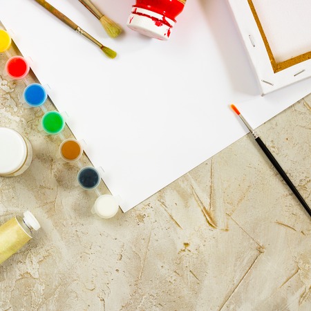 Flatlay with bright water colour paints in small round containers and bottles and palettes as frame, brush and white empty paper sheet in center on grey cement backcground, mess on artist deskの写真素材