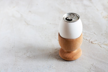 White egg in wooden egg cup, opened alluminium can as top of egg on grey cement textured background...