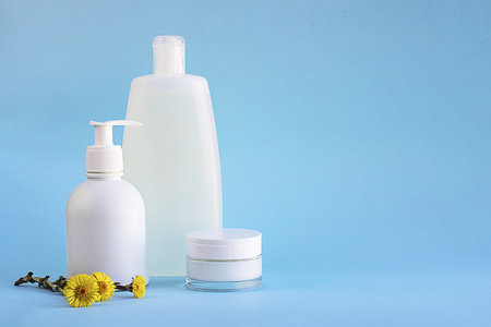 White plastic mockup bottles for shampoo, shower gel, dispenser, glass container for cream, flowers of healing herb foalfoot or Tussilago farfara on lighnt blue empty background with copy spaceの写真素材