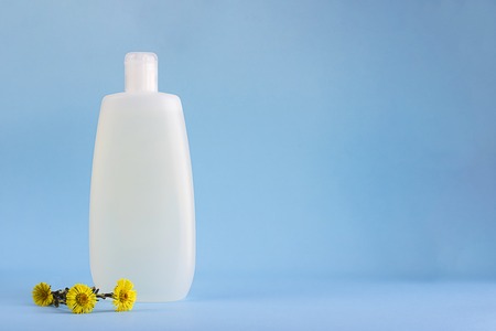 White plastic mockup bottle for shampoo or shower gel, dispenser, glass container for cream, flowers of healing herb foalfoot, Tussilago farfara on lighnt blue empty background with copy spaceの写真素材