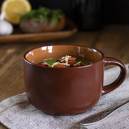 Green cream soup with red croutons and grated cheese in brown mug on natural ctoth on angle of old rustic wooden table, defocused ingredients. Square shot Healthy vegetarian foodの写真素材