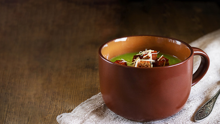 Green cream soup with red croutons with paprica, grated cheese in brown mug on natural ctoth on angle of old rustic wooden table Horizontal with copy space for text and design Healthy vegetarian foodの写真素材