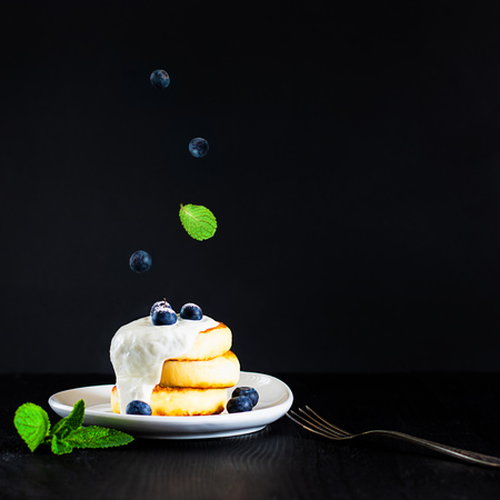 cottage cheese pancakes one on another topped with cream, blueberries and sugar powder on black background with fork and green mint leaves, breakfast, Square with copy space for text and designの写真素材
