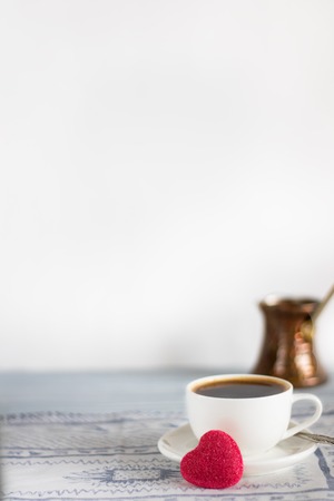 White cup of black coffee and heart shape bright pink candy on light background with defocused copper brewing pot, Vertical with copy space for text or designの写真素材