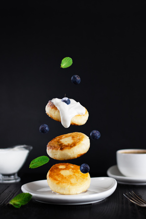 three cottage cheese pancakes topped with cream, blueberries, mint leaves fall down on white plate, defocused cup of coffee and saucer, black background, breakfast, Levitation Vertical copy spaceの写真素材