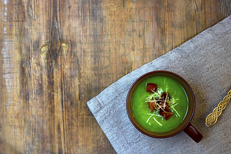 Green cream soup with red croutons and grated cheese in brown mug on natural ctoth on angle of old rustic wooden table. Horizontal with copy space. Healthy vegetarian food. Top view.の写真素材