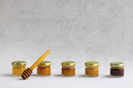 Five Small glass jar with metal cap with different kinds and colours of honey put in line and wooden spoon isolate and on grey cement background with copy space. Healthy product, natural. Horizontalの写真素材