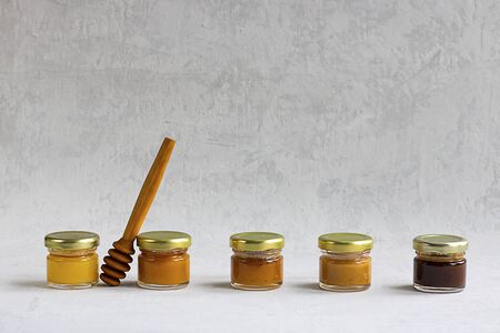 Five Small glass jar with metal cap with different kinds and colours of honey put in line and wooden spoon isolate and on grey cement background with copy space. Healthy product, natural. Horizontalの写真素材