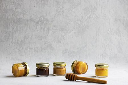 Five Small glass jar with metal cap with different kinds and colours of honey put in line and wooden spoon isolate and on grey cement background with copy space. Healthy product, natural. Horizontalの写真素材