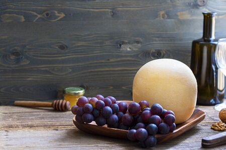 Round head of cheese Kostromskoy on textured dark wooden background on square plate with grapes, honey, defocused bottle and glass. Traditional product, Kostroma, Russia. Horizontal with copy space.の写真素材