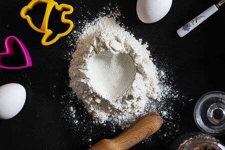 Handful of sieved flour with heart shape hole in the center on black table and things for baking around, St Valentines concept, Love relations, cosy home, Holiday, date, home made baking, Horizontalの写真素材