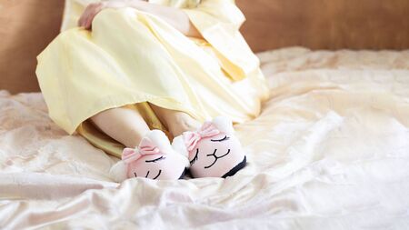 female feet wearing cute llama trendy pink slippers, lay on bed with wooden headboard and on pastel silk gown. Home Rest Relax Holidays Horizontal with copy spaceの写真素材