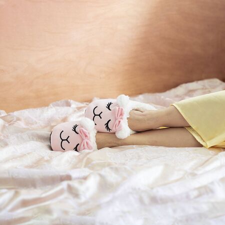 female feet wearing cute llama trendy pink slippers, lay on bed with wooden headboard and on pastel silk gown. Home Rest Relax Holidays Square with copy spaceの写真素材