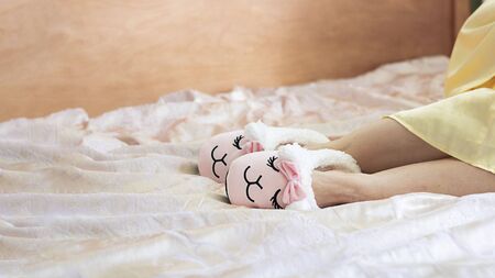 female feet wearing cute llama trendy pink slippers, lay on bed with wooden headboard and on pastel silk gown. Home Rest Relax Holidays Horizontal banner with copy spaceの写真素材