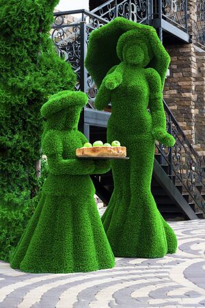 Green sculptures of young girl and a lady with umprella in long dress made from artificial grass. Girl holds wooden tray with fresh apples. Urban design, city architecture, park decoration. Vertical.の写真素材