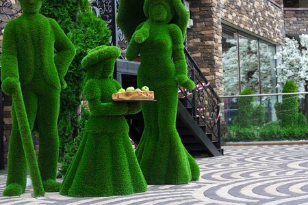 Green sculptures of young lady, man and woman with umprella made from artificial grass. Girl holds wooden tray with fresh apples. Urban design, city architecture, park decoration Horizontal copy spaceの写真素材