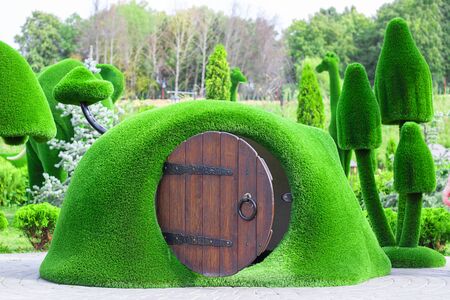 Green sculpture of small house with round wooden opened door surrounded by mushrooms made from artificial grass. Urban design, city architecture, park decoration Vertical with copy spaceの写真素材