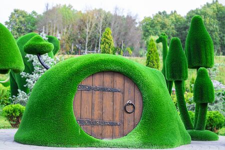 Green sculpture of small house with round wooden door surrounded by mushrooms made from artificial grass. Urban design, city architecture, park decoration Vertical with copy spaceの写真素材