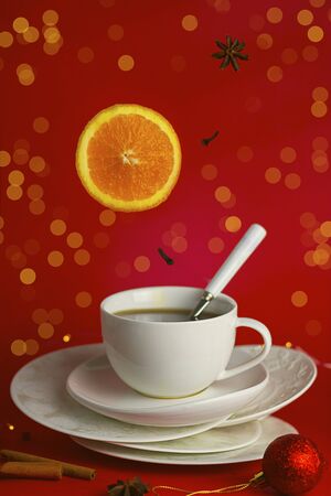 Steaming cup of tea with mockup spoon on the pile of saucers with falling ingredients orange and spicies on red background with hokeh. Christmas and new year concept. Vertical. Food levitationの写真素材