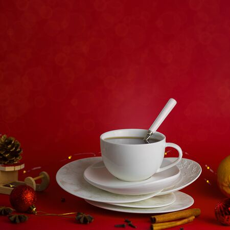 White steaming cup of hot tea or coffee with teaspoon with white mockup handle on the pile of saucers on red background with christmas decoration and spicies around. Square with copy space and bokehの写真素材