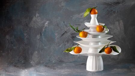 White dishes Pyramid like Christmas tree with mandarins with leaves on top, bunch below on dark Creative concept, dishware, veg, agriculture, vegetable, kitchen, restaurant, postcard, Xmas, New Yearの写真素材