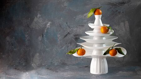 White dishes Pyramid like Christmas tree with mandarins with leaves on top, bunch below on dark Creative concept, dishware, veg, agriculture, vegetable, kitchen, restaurant, postcard, Xmas, New Yearの写真素材