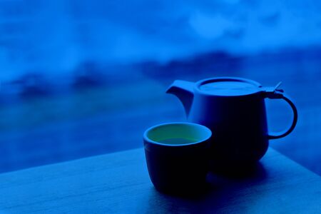Minimal style set with ceramic tea pot and cup with tea on the table at cafe on the background of defocused rainy street. Horizontal with copy space. Classic, blue monochrome, trend 2020の写真素材