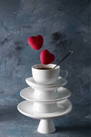 White saucer pyramid cup of tea with spoon on top. With two candy red hearts flying over Dark background. Creative concept St. Valentine's day dishware, love, relations, gift, celebrate, restaurant,の写真素材