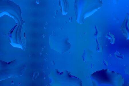 Abstract water drops on glass with defocused background. Classic, blue monochrome, trend 2020. Horizontal. Rainy day. View through the window.の写真素材