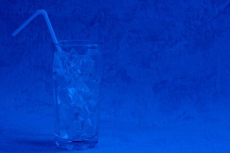 Tall empty glass with ice cubes and plastic straw ready to pour cold summer beverage on cement background with copy space. Classic, blue monochrome, trend 2020の写真素材