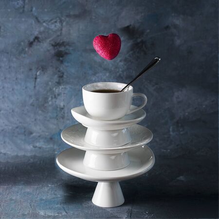White saucer pyramid with cup of tea on top and flying red hearts candy over it. Creative concept, love, relations, gift, celebrate, restaurant, food levitation, St. Valentine's day. Dark background.の写真素材