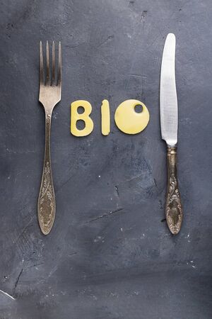 Food concept, letters BIO made of cheese with holes between vintage fork and knife on empty grey background. Vertical. Creative for dairy, diet, health, menu, organic product.の写真素材