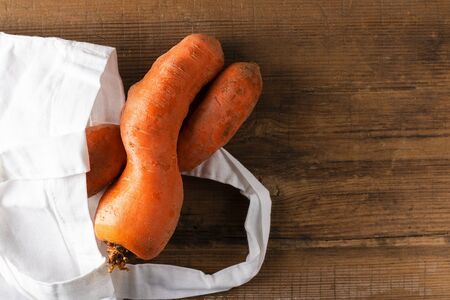 Curved ugly carrots in white eco bag on brown old wooden background with copy space. Vegetables with unusual strange shape. Horizontal. Food that grocery store, supermarket usually throw away. Reuseの写真素材