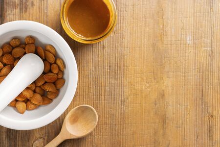 Glass jar with apricot kernel butter, spoon, white mortar with pestle with whole nuts and spoon on wooden background with copy space.の写真素材