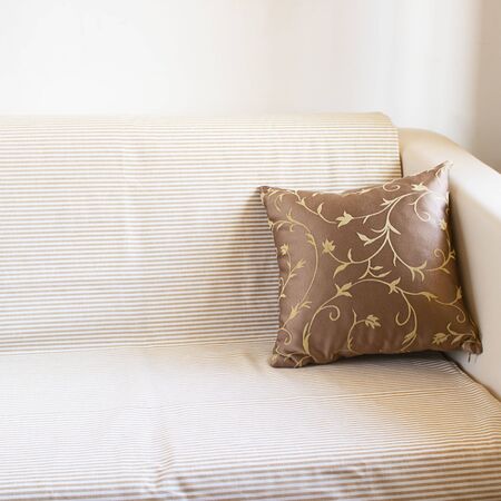 Beautiful brown pillow on light sofa covered by linen blancet with pale stripes on the background of white wall. Square with copy space. Decoration in living room interior.の写真素材