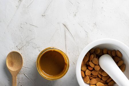 Glass jar with aprikot kernel butter, wooden spoon, white mortar with pestle with whole nuts and spoon on textured cement background with copy space. Product for cardio vascular system, vitamims.の写真素材