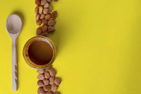 Glass jar with aprikot kernel butter, wooden spoon with whole nuts on yellow background, copy space. Product for cardio vascular systemの写真素材